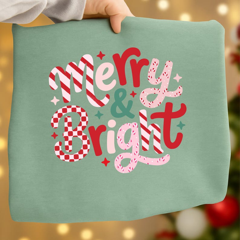 以下が含まれることがあります： 「Merry & Bright」の文字がデザインされた、明るい緑色のクッション。赤、ピンク、白のストライプとチェック柄で、星とドットのアクセントが施されています。クッションは人が持っています。