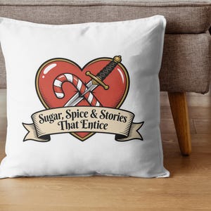 May include: White throw pillow with a heart-shaped design featuring a candy cane, sword, and banner. The banner reads "Sugar, Spice & Stories That Entice". The design has red, gold, and black accents. The pillow is on a wooden surface.