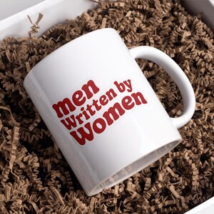 May include: White ceramic mug with the text "men Written by Women" in red lettering. The mug is inside a white box filled with brown crinkle paper. The mug has a curved handle.