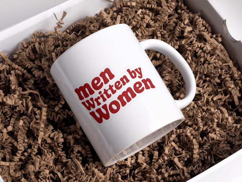 May include: White ceramic mug with the text "men Written by Women" in red lettering. The mug is inside a white box filled with brown crinkle paper. The mug has a curved handle.