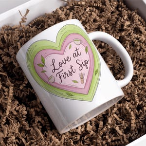 Puede incluir: Taza de cer&aacute;mica blanca con un dise&ntilde;o en forma de coraz&oacute;n en tonos rosa y verde, con el texto "Love at First Sip". La taza est&aacute; en una caja blanca llena de papel de relleno marr&oacute;n.