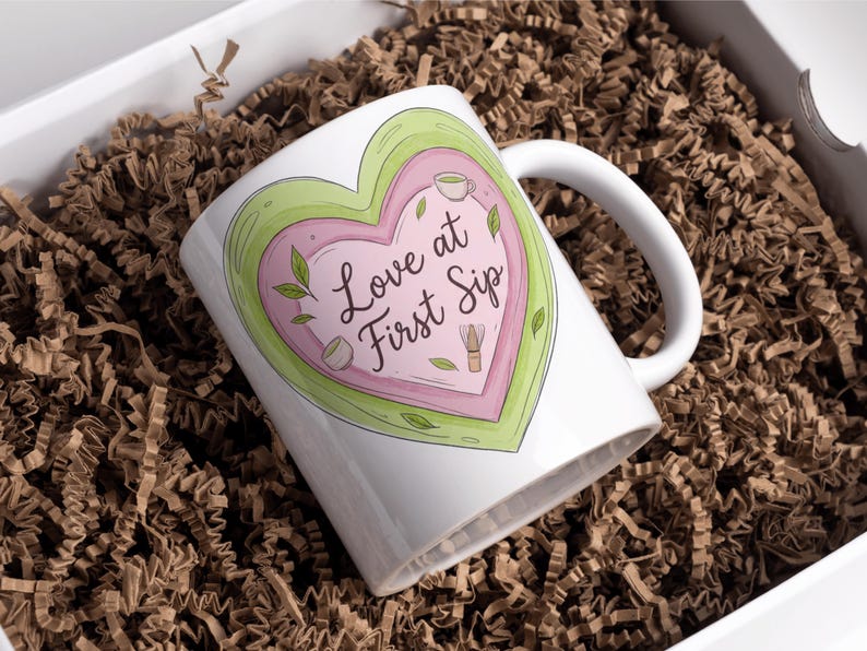 Puede incluir: Taza de cer&aacute;mica blanca con un dise&ntilde;o en forma de coraz&oacute;n en tonos rosa y verde, con el texto "Love at First Sip". La taza est&aacute; en una caja blanca llena de papel de relleno marr&oacute;n.