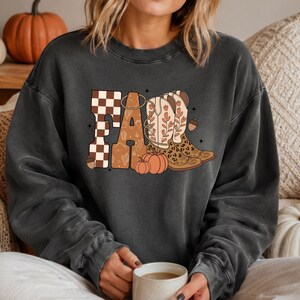 May include: A charcoal gray crewneck sweatshirt featuring a graphic print of the word FALL. The letters incorporate a brown and white checkerboard pattern, a desert landscape, and a pair of western boots with floral and leopard print details.