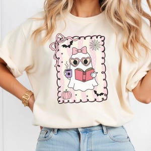 May include: Cream-colored t-shirt featuring a cartoon ghost reading a book, wearing heart-shaped glasses and a bow. The ghost is holding a cup of coffee. The design includes bats, a spiderweb, and a pumpkin, all within a pink and white striped frame.