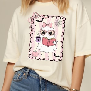 May include: Cream-colored t-shirt featuring a graphic of a ghost reading a book, wearing heart-shaped glasses and a bow. The ghost is holding a coffee cup. The graphic is framed with a pink border, bats, a spiderweb, and a pumpkin.