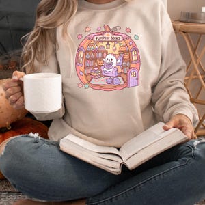 May include: Beige sweatshirt featuring a whimsical illustration of a pumpkin-shaped library with the text "PUMPKIN BOOKS". The design includes a ghost reading, a cat, books, and autumn leaves. The person in the image is holding a white mug and a book.