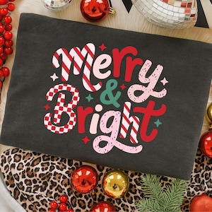 以下が含まれることがあります： 「Merry & Bright」の文字がカラフルで華やかなデザインでプリントされたダークグレーのスウェットシャツ。赤、ピンク、白のキャンディケーンストライプと市松模様が特徴です。スウェットシャツはクリスマスオーナメントに囲まれています。