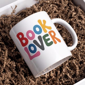 May include: White ceramic mug with the text "BOOK LOVER" in colorful, playful letters. The mug is inside a white box filled with brown crinkle paper, suggesting a gift or presentation.