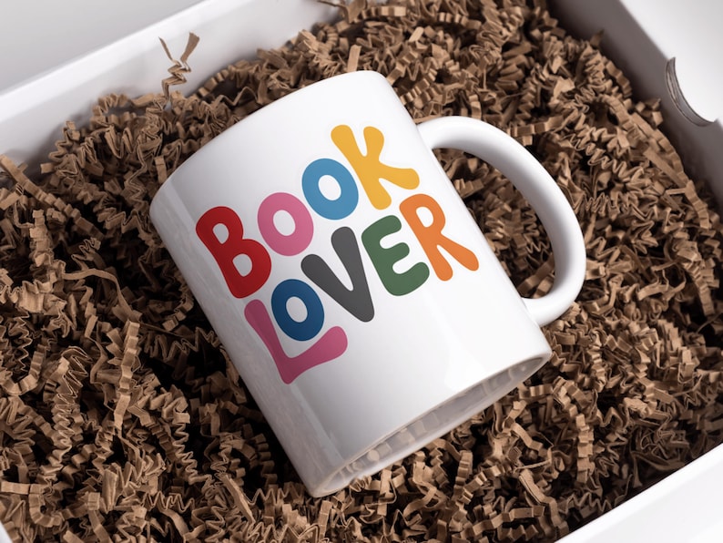 May include: White ceramic mug with the text "BOOK LOVER" in colorful, playful letters. The mug is inside a white box filled with brown crinkle paper, suggesting a gift or presentation.