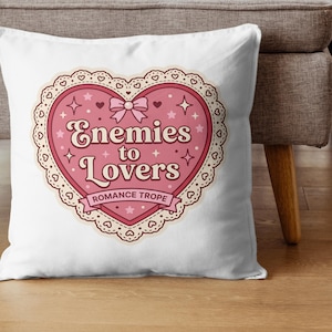 May include: White throw pillow with a pink heart-shaped design. The design features the text "Enemies to Lovers" with a bow, stars, and a lace-like border. The words "Romance Trope" are also included.