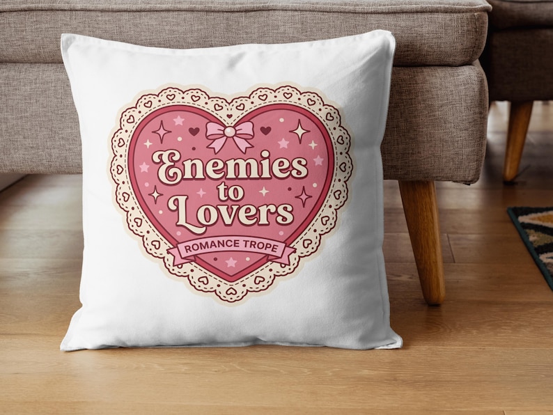 May include: White throw pillow with a pink heart-shaped design. The design features the text "Enemies to Lovers" with a bow, stars, and a lace-like border. The words "Romance Trope" are also included.