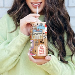 May include: A clear glass tumbler with a bamboo lid and glass straw, filled with iced coffee. The exterior features colorful stickers including a newspaper with the text Stop stressing over shitty people and a sign reading Bad vibes don't go with my lipstick.