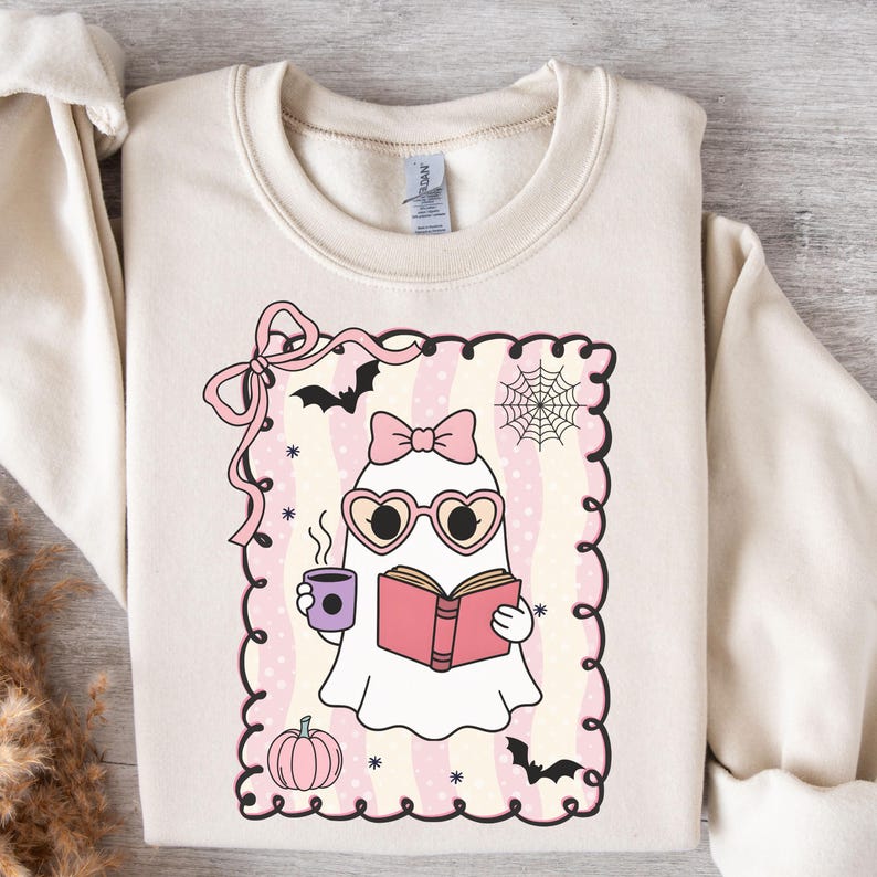 May include: Cream-colored sweatshirt featuring a cute ghost illustration. The ghost wears heart-shaped glasses and a bow, holding a book and a coffee cup. The design includes bats, a spiderweb, a pumpkin, and a pink bow.