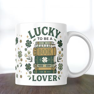 May include: White ceramic mug with a green and gold design. The mug features the text "LUCKY TO BE A BOOK LOVER" with illustrations of books, clovers, and flowers. The mug has a curved handle.