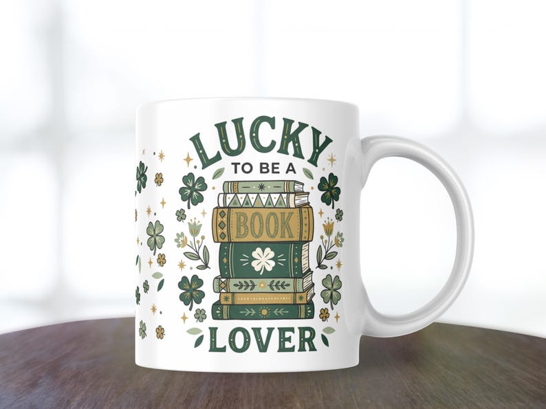 May include: White ceramic mug with a green and gold design. The mug features the text "LUCKY TO BE A BOOK LOVER" with illustrations of books, clovers, and flowers. The mug has a curved handle.