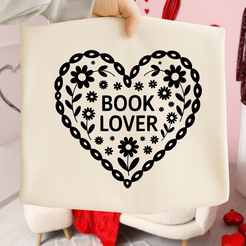 Puede incluir: Bolso tote color crema con un dise&ntilde;o en forma de coraz&oacute;n negro lleno de flores y las palabras "BOOK LOVER". El coraz&oacute;n est&aacute; delineado con un borde decorativo. El bolso lo sostiene una persona.
