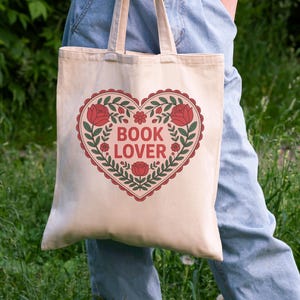 May include: A beige tote bag with a heart-shaped design. The heart is outlined in red with a scalloped edge and filled with green leaves, red roses, and the words "BOOK LOVER" in red letters. The bag has beige handles.