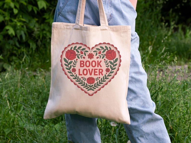 May include: A beige tote bag with a heart-shaped design. The heart is outlined in red with a scalloped edge and filled with green leaves, red roses, and the words "BOOK LOVER" in red letters. The bag has beige handles.