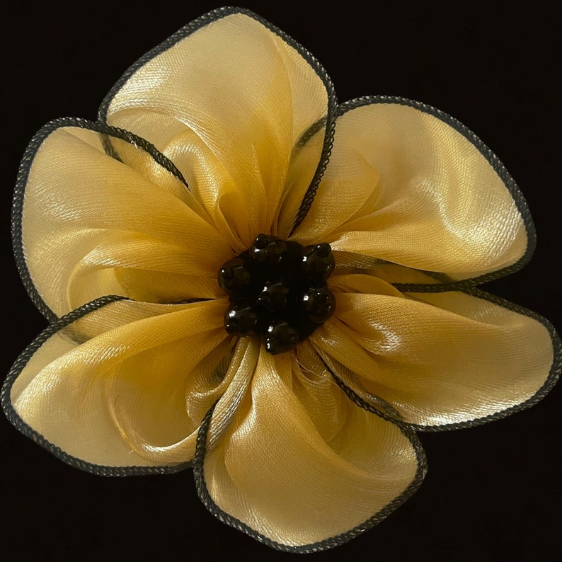 Puede incluir: Una gran flor amarilla con detalles de cuentas negras. La flor est&aacute; hecha de tela transparente y tiene un aspecto arrugado.