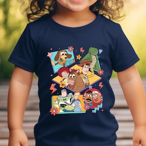 May include: Navy blue t-shirt featuring a colorful graphic of characters from the animated film "Toy Story." The design includes images of Woody, Buzz Lightyear, and other characters. The shirt is suitable for children.