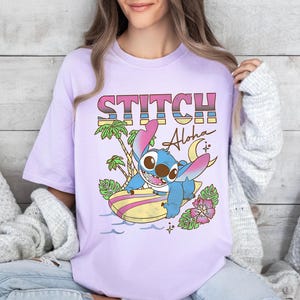 Puede incluir: Camiseta lila claro con la palabra "STITCH" en una fuente de arco&iacute;ris y un personaje de dibujos animados surfeando en una tabla de surf. La camiseta tambi&eacute;n incluye el texto "Aloha".