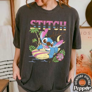 Puede incluir: Camiseta gris oscuro con un gr&aacute;fico de Stitch surfeando, la palabra "STITCH" arriba. El dise&ntilde;o incluye palmeras, olas y una luna creciente. La camiseta es de la marca Comfort Colors.