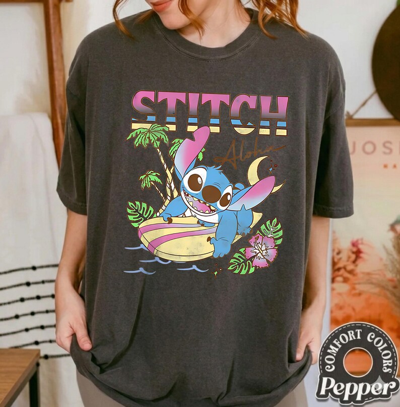 Puede incluir: Camiseta gris oscuro con un gr&aacute;fico de Stitch surfeando, la palabra "STITCH" arriba. El dise&ntilde;o incluye palmeras, olas y una luna creciente. La camiseta es de la marca Comfort Colors.