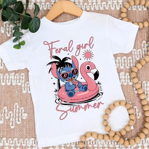 Pu&ograve; includere: Maglietta bianca con un personaggio dei cartoni animati blu con occhiali da sole su un galleggiante a forma di fenicottero rosa. Il testo "Feral girl summer" &egrave; scritto in rosa.