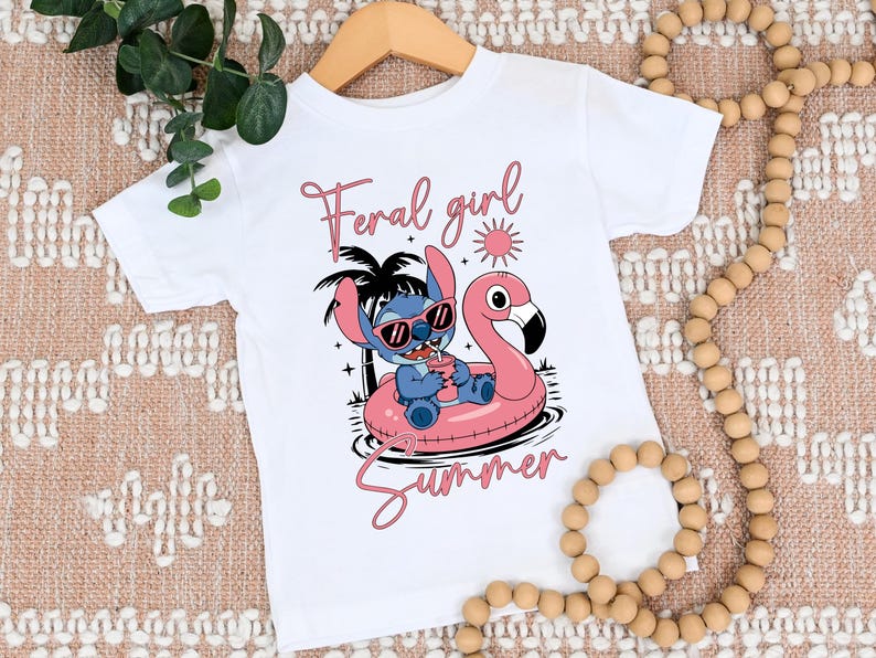 Pu&ograve; includere: Maglietta bianca con un personaggio dei cartoni animati blu con occhiali da sole su un galleggiante a forma di fenicottero rosa. Il testo "Feral girl summer" &egrave; scritto in rosa.