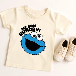 May include: Cream-colored toddler t-shirt with a blue Cookie Monster graphic and the text "ME SON HUNGRY!". A pair of cream-colored canvas sneakers are next to the shirt.
