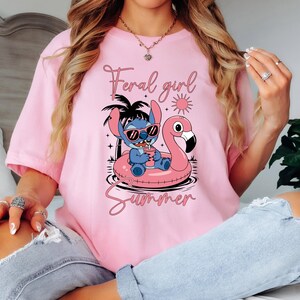 Pu&ograve; includere: T-shirt rosa chiaro con un personaggio dei cartoni animati Stitch con occhiali da sole su un galleggiante a forma di fenicottero rosa. Il testo "Feral girl Summer" &egrave; stampato in corsivo. La maglietta &egrave; a maniche corte.