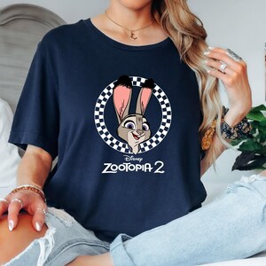 May include: Navy blue t-shirt featuring a cartoon image of Judy Hopps from Disney's Zootopia 2. The image is in a circular frame with a black and white checkered pattern. The text "Zootopia 2" is below the image.