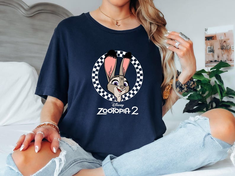 May include: Navy blue t-shirt featuring a cartoon image of Judy Hopps from Disney's Zootopia 2. The image is in a circular frame with a black and white checkered pattern. The text "Zootopia 2" is below the image.