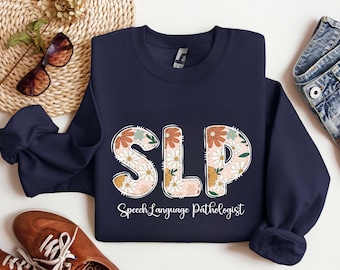 May include: Navy blue sweatshirt with the letters "SLP" in a floral design. The text "Speech Language Pathologist" is below the letters. The sweatshirt is displayed with sunglasses, a woven mat, and brown shoes.