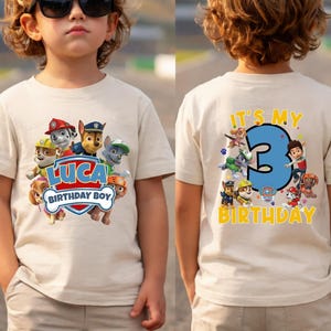 May include: A beige short-sleeve t-shirt featuring Paw Patrol characters. The front displays the name Luca and the text Birthday Boy. The back features a large blue number 3 with the text It's My Birthday and additional character graphics.