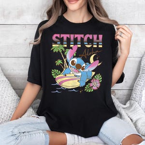 Puede incluir: Camiseta negra con un gr&aacute;fico colorido de Stitch surfeando en una tabla de surf. El dise&ntilde;o incluye la palabra "Stitch" en una fuente retro, palmeras y la palabra "Aloha".