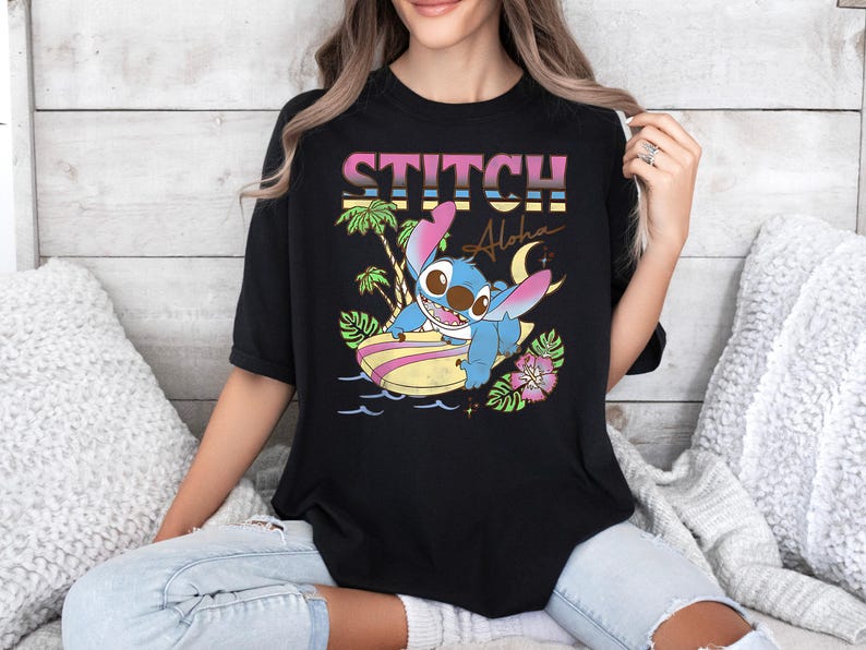 Puede incluir: Camiseta negra con un gr&aacute;fico colorido de Stitch surfeando en una tabla de surf. El dise&ntilde;o incluye la palabra "Stitch" en una fuente retro, palmeras y la palabra "Aloha".