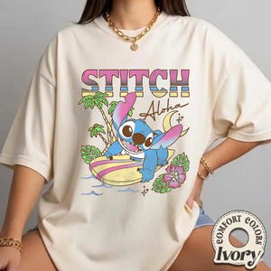 Puede incluir: Camiseta color marfil con un dise&ntilde;o de Stitch surfeando y el texto "STITCH Aloha". El dise&ntilde;o incluye palmeras, una luna creciente y flores tropicales. La camiseta es de Comfort Colors.