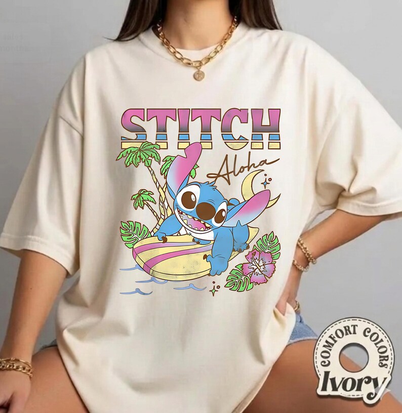 Puede incluir: Camiseta color marfil con un dise&ntilde;o de Stitch surfeando y el texto "STITCH Aloha". El dise&ntilde;o incluye palmeras, una luna creciente y flores tropicales. La camiseta es de Comfort Colors.