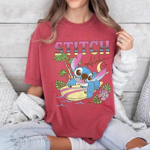 Puede incluir: Camiseta roja con un dise&ntilde;o de Stitch de Lilo & Stitch. El dise&ntilde;o incluye la palabra "STITCH" en letras retro coloridas, junto con Stitch en una tabla de surf, palmeras y la palabra "Aloha".