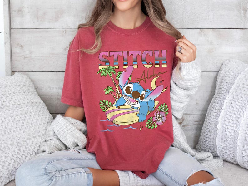 Puede incluir: Camiseta roja con un dise&ntilde;o de Stitch de Lilo & Stitch. El dise&ntilde;o incluye la palabra "STITCH" en letras retro coloridas, junto con Stitch en una tabla de surf, palmeras y la palabra "Aloha".