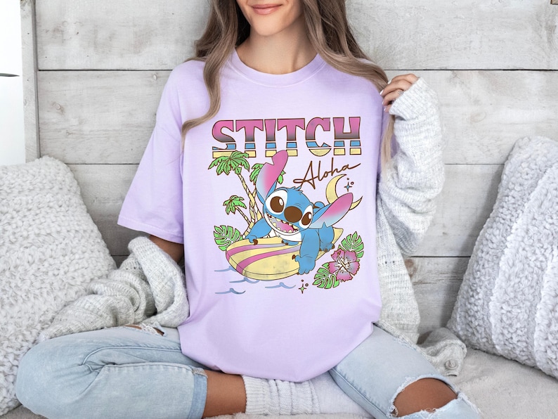 Puede incluir: Camiseta lila claro con la palabra "STITCH" en una fuente de arco&iacute;ris y un personaje de dibujos animados surfeando en una tabla de surf. La camiseta tambi&eacute;n incluye el texto "Aloha".