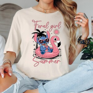 Pu&ograve; includere: T-shirt color crema con un personaggio dei cartoni animati con occhiali da sole su un galleggiante a forma di fenicottero rosa. Il testo "Feral girl Summer" &egrave; scritto in corsivo rosa sopra e sotto la grafica. Design casual a tema estivo.