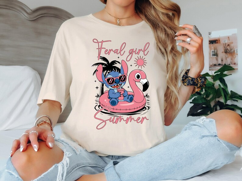 Pu&ograve; includere: T-shirt color crema con un personaggio dei cartoni animati con occhiali da sole su un galleggiante a forma di fenicottero rosa. Il testo "Feral girl Summer" &egrave; scritto in corsivo rosa sopra e sotto la grafica. Design casual a tema estivo.