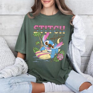 Puede incluir: Camiseta verde oliva con la palabra "STITCH" en letras de bloque rosas y amarillas sobre un personaje de dibujos animados en una tabla de surf. El gr&aacute;fico incluye palmeras, una luna creciente y la palabra "Aloha".