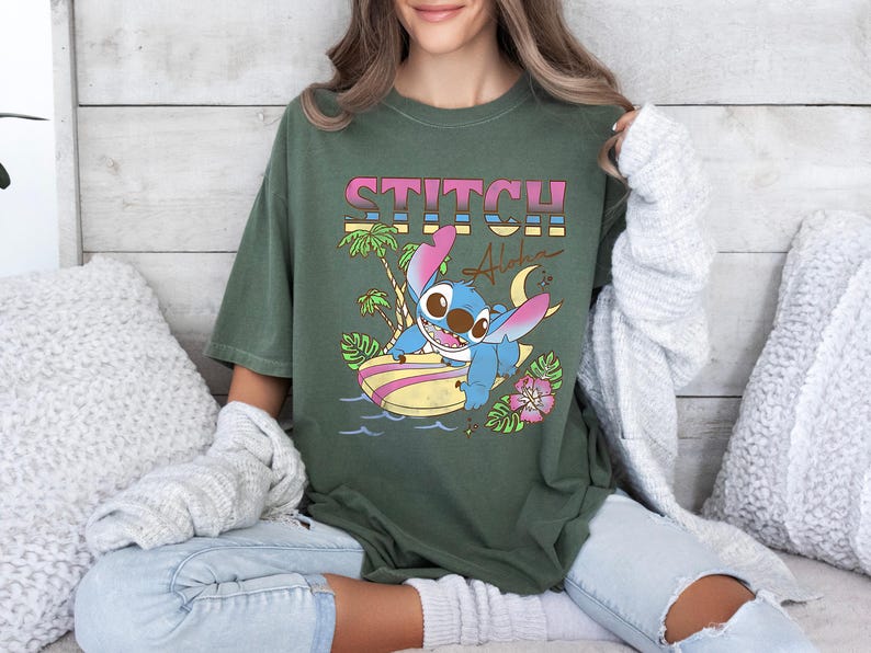 Puede incluir: Camiseta verde oliva con la palabra "STITCH" en letras de bloque rosas y amarillas sobre un personaje de dibujos animados en una tabla de surf. El gr&aacute;fico incluye palmeras, una luna creciente y la palabra "Aloha".