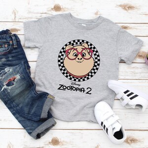 May include: A gray t-shirt featuring a cartoon character from Zootopia 2 wearing pink glasses, set within a black and white checkered circle. The shirt is paired with blue jeans and white sneakers with black stripes.