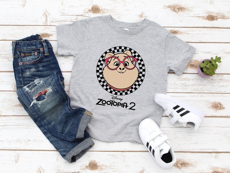 May include: A gray t-shirt featuring a cartoon character from Zootopia 2 wearing pink glasses, set within a black and white checkered circle. The shirt is paired with blue jeans and white sneakers with black stripes.