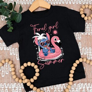 Pu&ograve; includere: T-shirt nero con un personaggio dei cartoni animati con occhiali da sole su un galleggiante a forma di fenicottero rosa. La maglietta mostra le parole "Feral girl Summer" in corsivo rosa. Il design include un sole e palme.