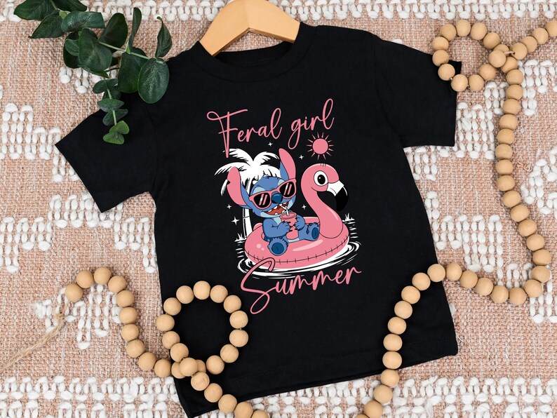 Pu&ograve; includere: T-shirt nero con un personaggio dei cartoni animati con occhiali da sole su un galleggiante a forma di fenicottero rosa. La maglietta mostra le parole "Feral girl Summer" in corsivo rosa. Il design include un sole e palme.